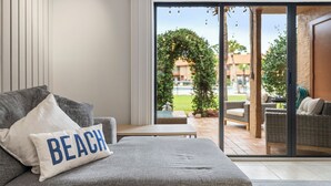 A modern living room with a grey sofa, a pillow with "BEACH" text, and a view of an outdoor patio area with wicker furniture and greenery. - Steps to Beach - 2 MBR-2.5 Bath, Sleeps 6, Gulf Highlands Beach Resort Townhome (Panama City Beach)