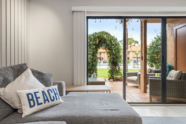 A modern living room with a grey sofa, a pillow with "BEACH" text, and a view of an outdoor patio area with wicker furniture and greenery.