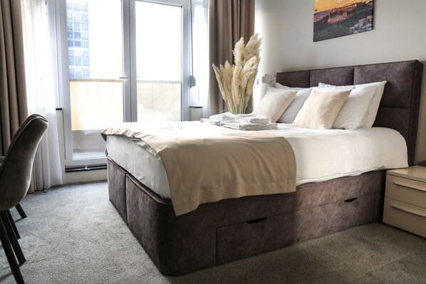 A modern bedroom with a large bed, a bedside table, a chair, and a view of the outside. - Smart Apartments Templum (Belgrade)