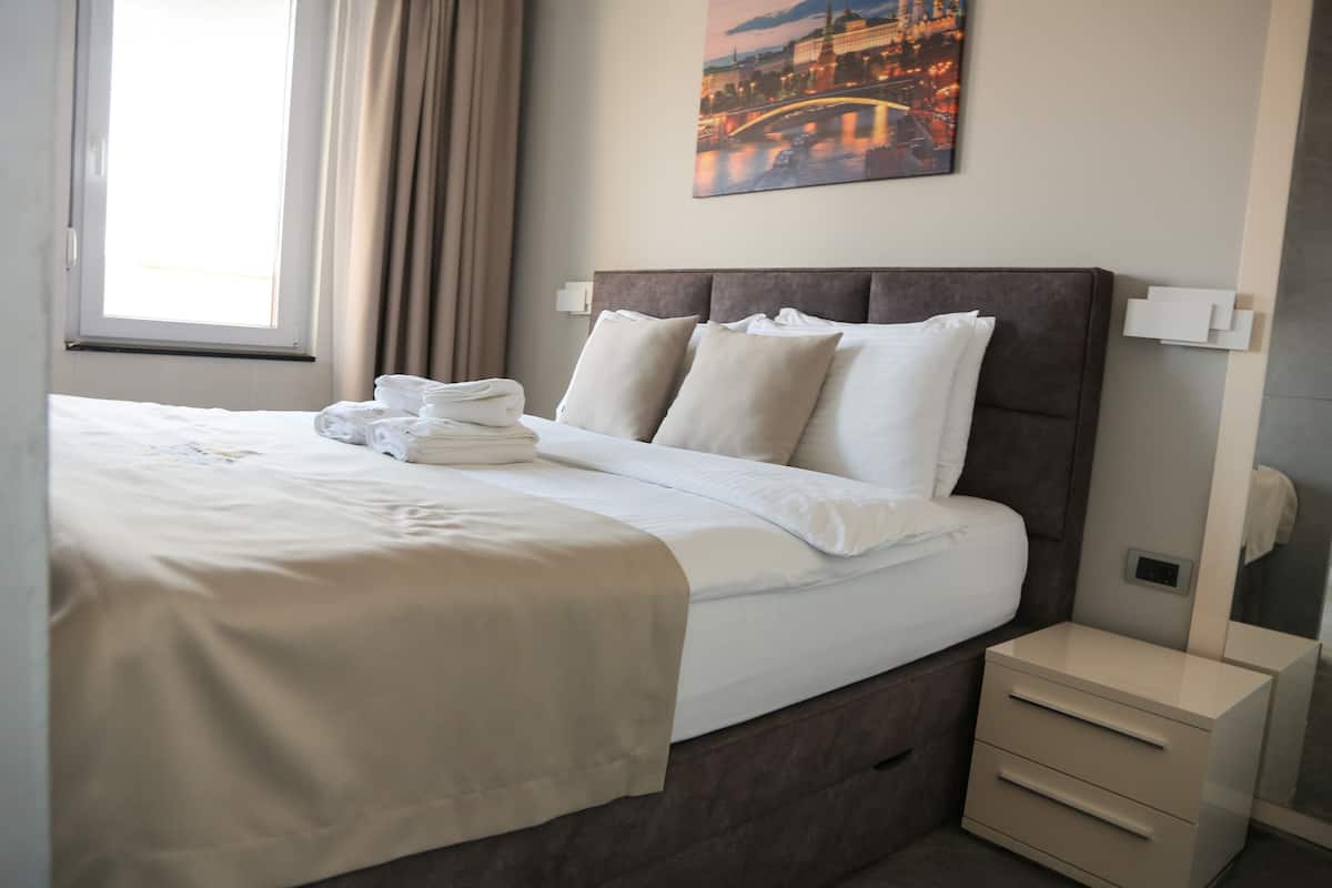 Ein ordentlich bezogenes Bett mit weißen Leinen und Kissen, ein Nachttisch mit einer Lampe und ein Stadtbild an der Wand.