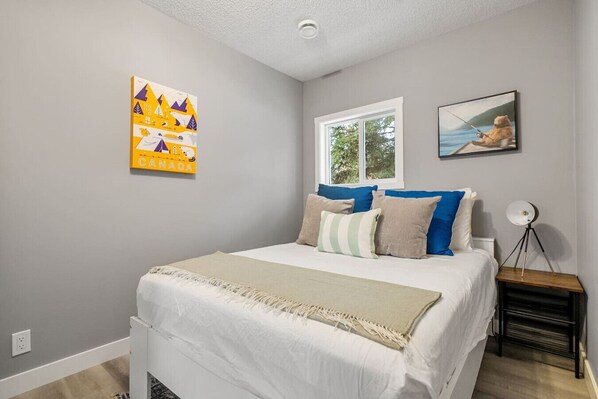 A bedroom with a bed, pillows, a window, a painting, and a lamp on a bedside table.