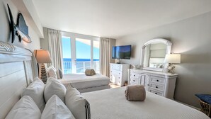 A hotel room with two beds, a television, a dresser, and a balcony with a view of the sea.