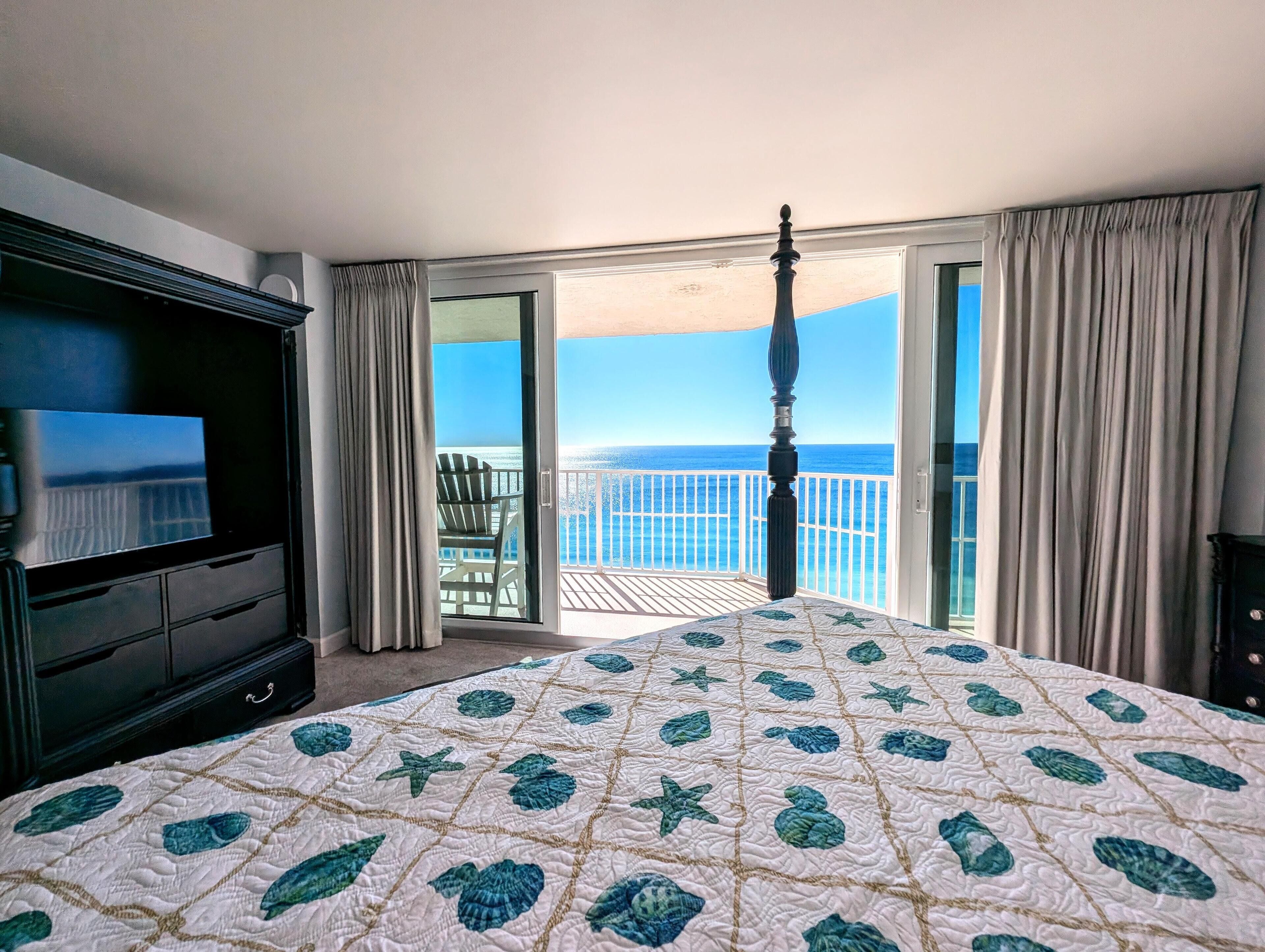 A hotel room with a bed, a TV, a balcony with a view of the ocean, and a chair.
