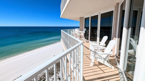 A balcony with white chairs and a view of the beach and ocean.