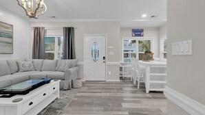 A modern living room with a sectional sofa, a dining area with white furniture, and a kitchen visible through a glass door. - Ocean Lakes Walk to Beach! Waterpark! Arcade! Dogs Welcome! (Myrtle Beach)