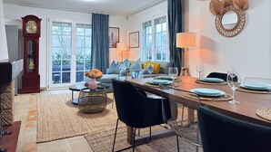 A dining area with a wooden table set for four, a black chair, and a round woven stool. A large mirror with a woven frame hangs on the wall, and a tall wooden clock is visible on the left. - Spacious 4 bedrooms House (Strassen)