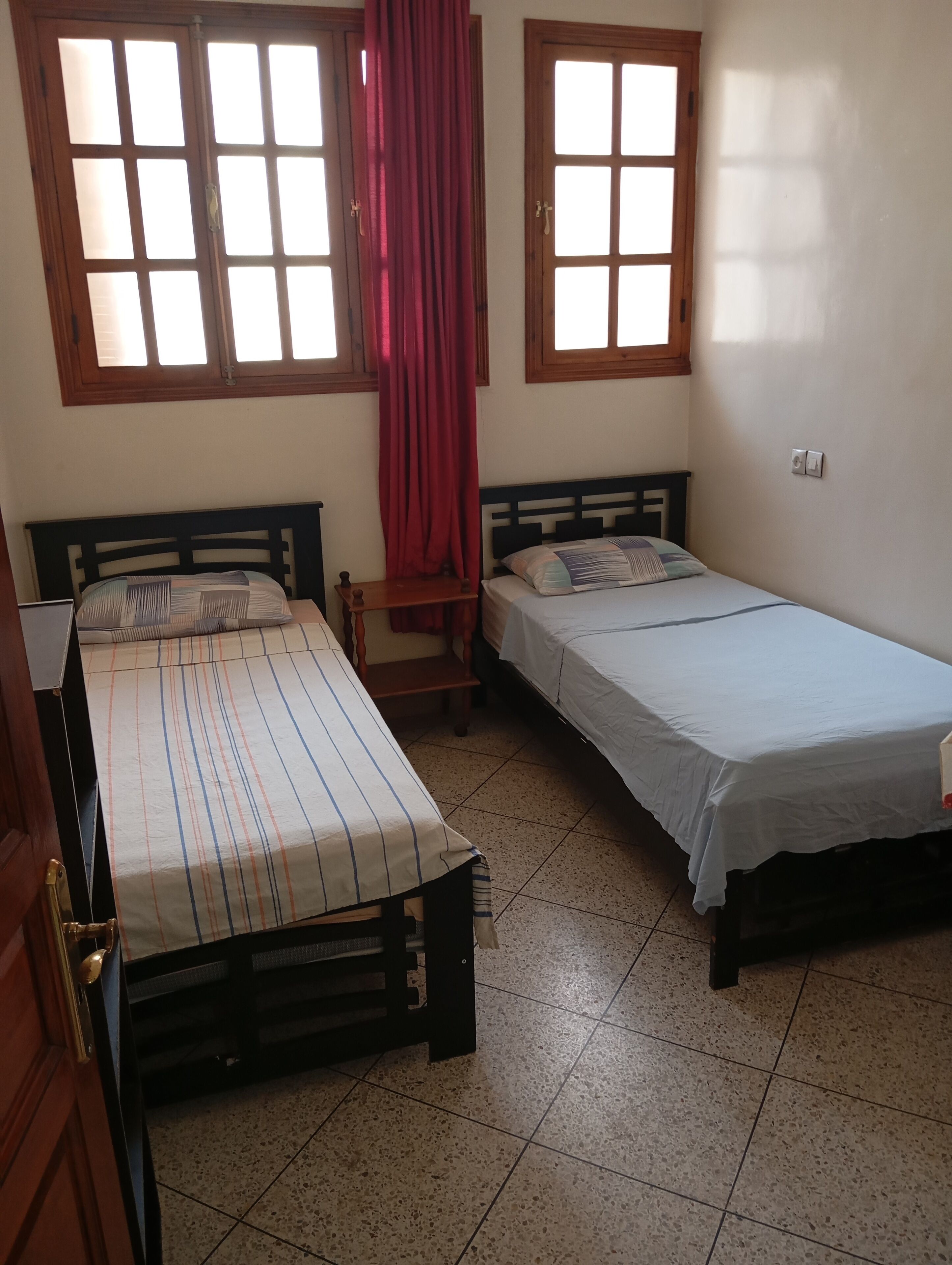 Une chambre avec deux lits simples, un banc en bois, un rideau rouge et deux fenêtres avec des cadres en bois.