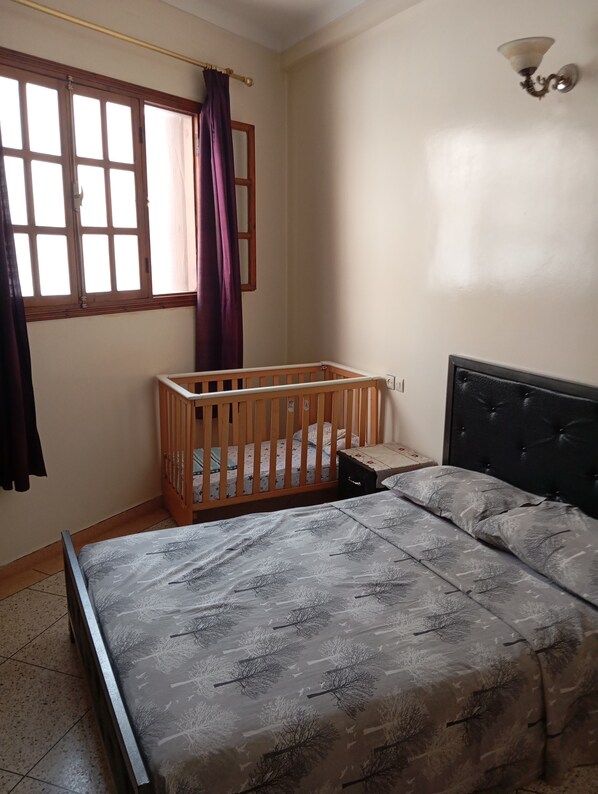 Une chambre à coucher avec un lit, un berceau, une fenêtre avec des rideaux et un luminaire fixé au mur.