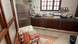 Eine Küche mit Kühlschrank, Herd, Spüle und diversen Küchenutensilien. Ein Fenster mit Jalousien, ein Warmwasserbereiter und eine Arbeitsfläche mit Geschirr und einem Mülleimer sind ebenfalls vorhanden.