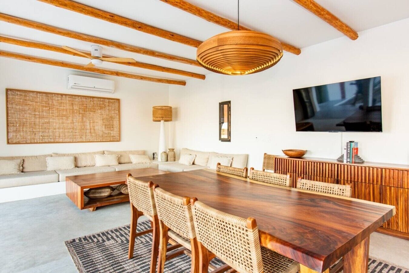A modern living room with a wooden dining table, woven chairs, a large woven wall hanging, a flat-screen TV, and a sofa with cushions.