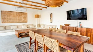Ein modernes Wohnzimmer mit einem Holzesstisch, gewobenen Stühlen, einem großen Wandbehang aus Gewebe, einem Flachbildfernseher und einer Couch mit Kissen.