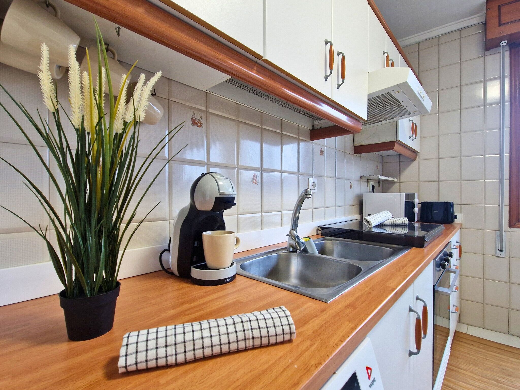 Une cuisine moderne avec des meubles blancs, un plan de travail en bois, une machine à café, un évier et une plante en pot.