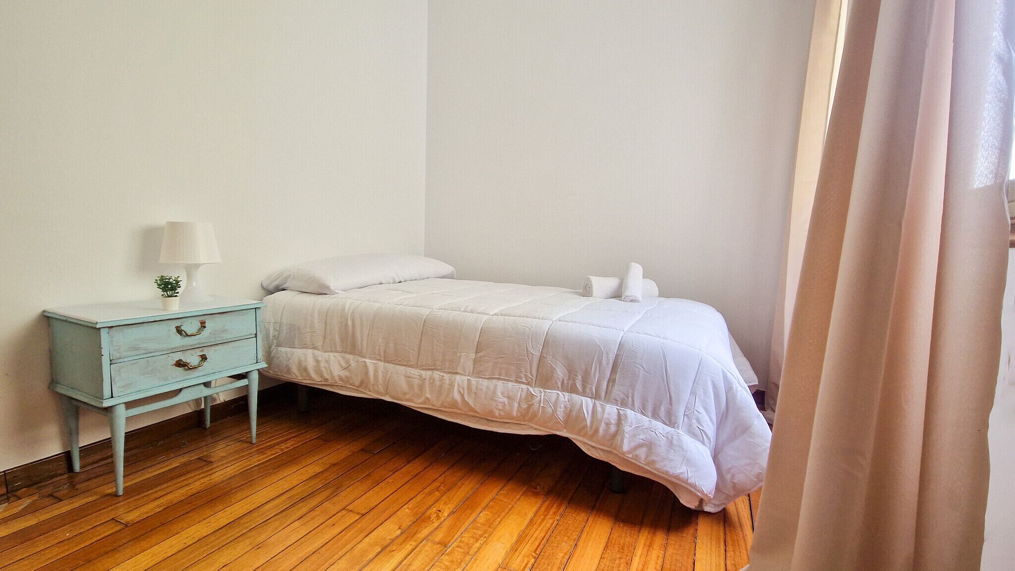 Una piccola camera con un letto singolo, pavimento in legno, una cassettiera azzurra con una lampada e una finestra con tende.