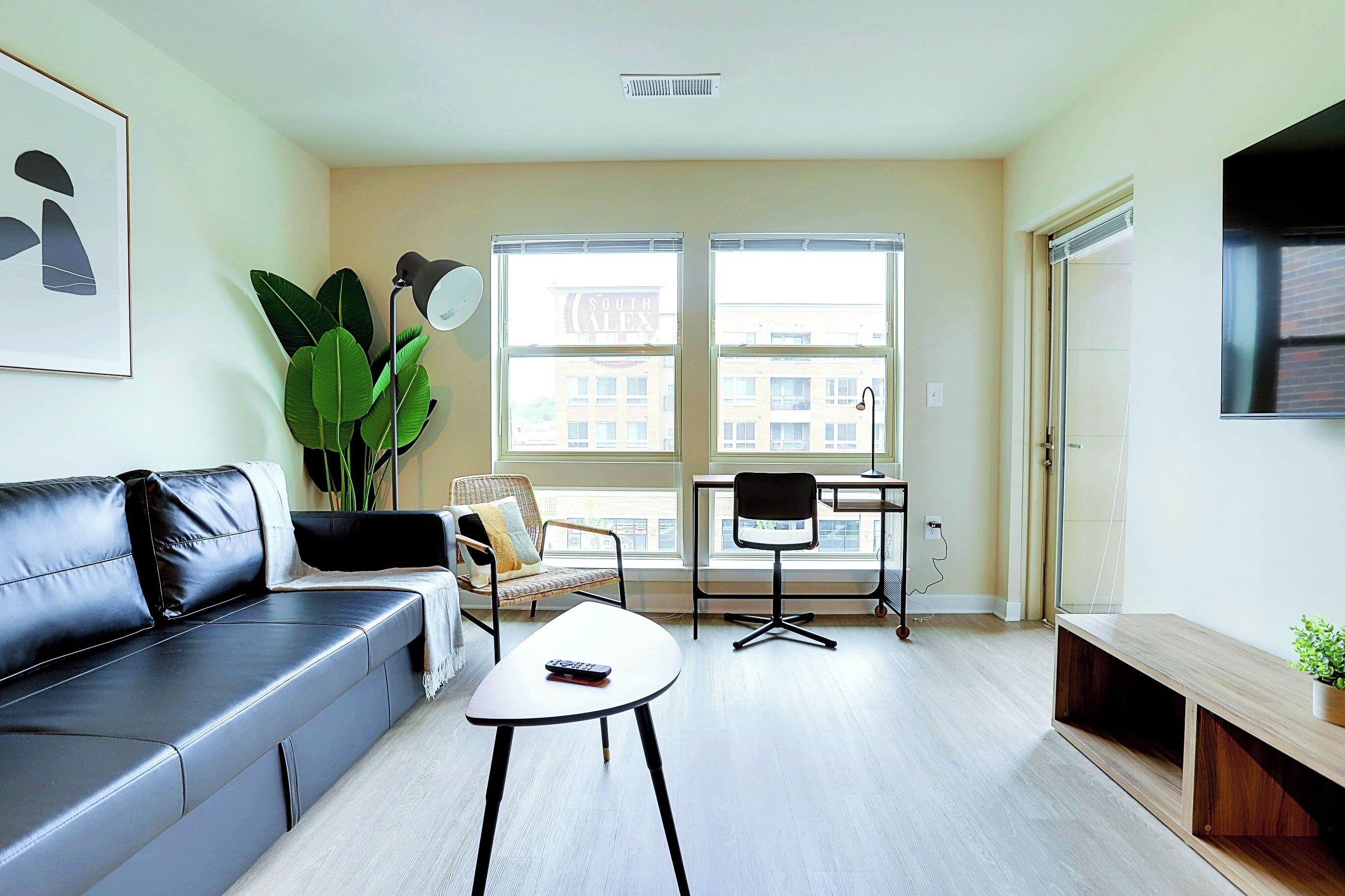 A modern living room with a black leather sofa, a white coffee table, a black office chair, and a wooden bench. Large windows provide a view of the city.