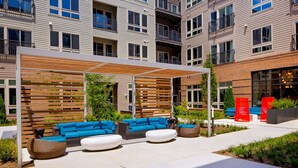 A modern outdoor seating area with blue sofas and white round ottomans, surrounded by a wooden pergola and greenery. - Charming DC Area Apartments (Alexandria)