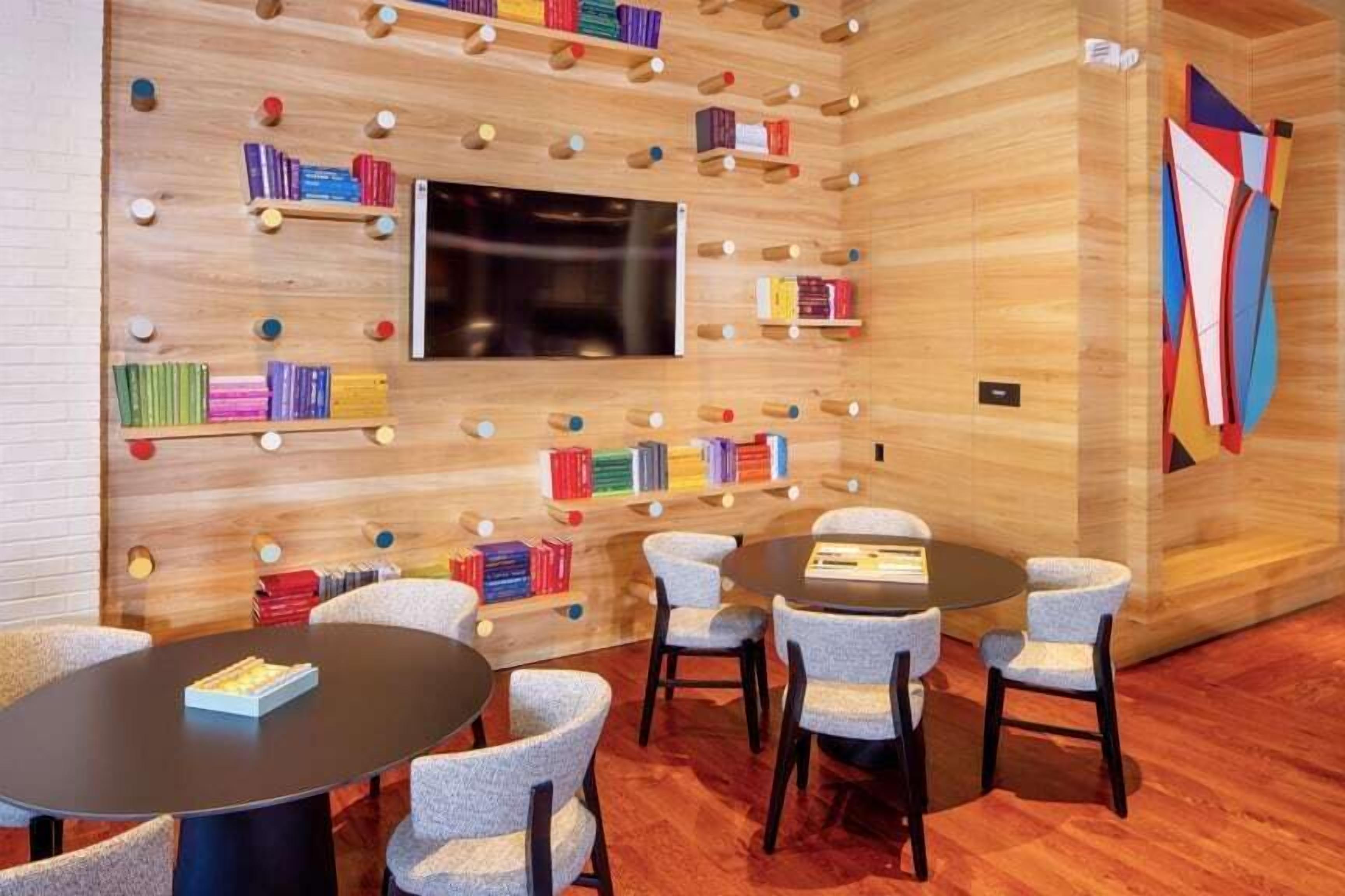 A modern room with a wooden wall, bookshelves, a flat-screen TV, and round tables with chairs.