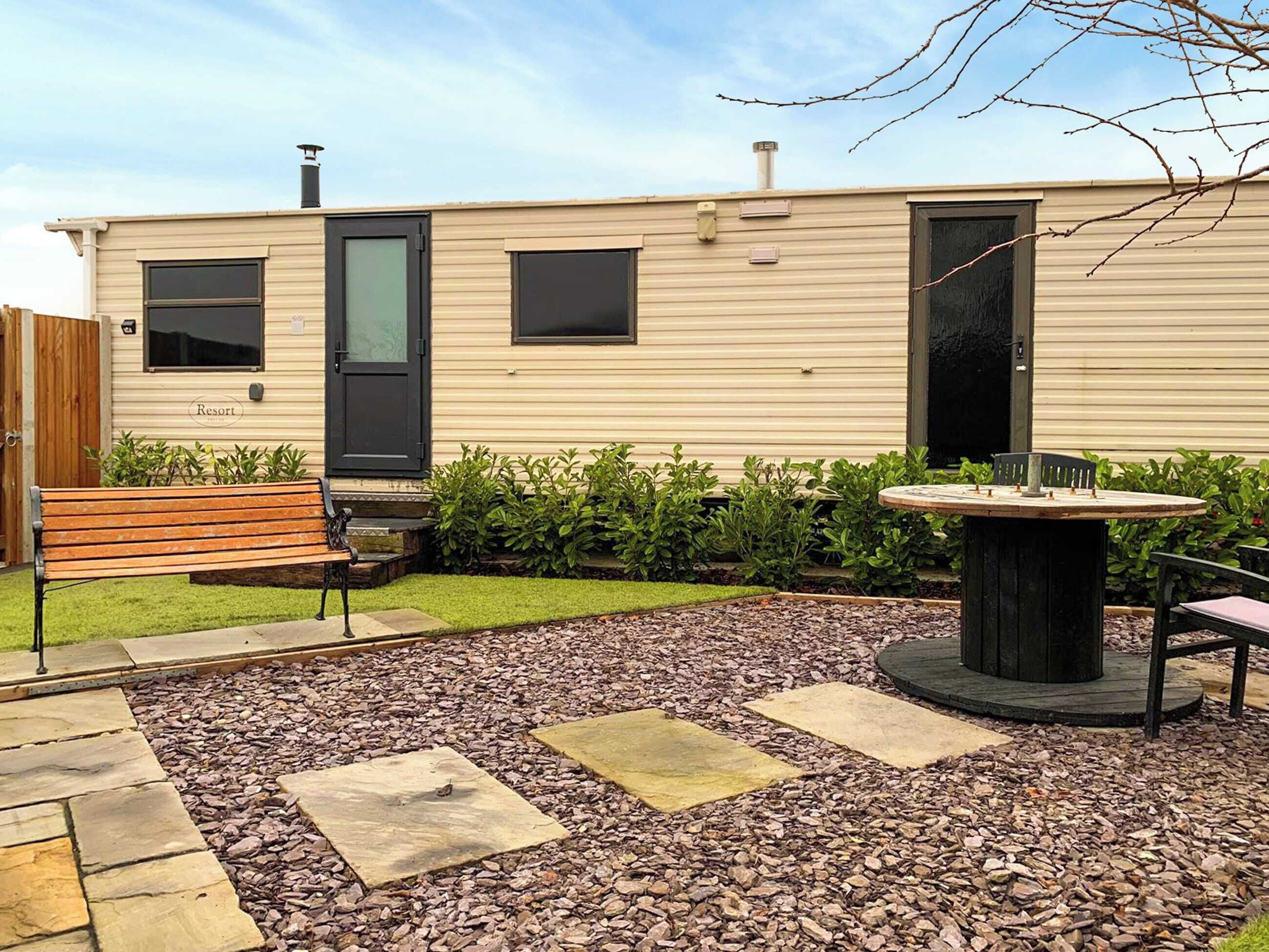 A mobile home with a patio area featuring a bench, a table, and a small garden with shrubs and a gravel path.