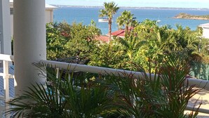 A balcony with a view of the sea, palm trees, and a building with a red roof. - Master suite + breakfast (saint martin)