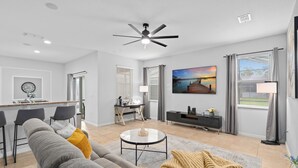A modern living room with a grey sofa, a coffee table, and a TV. There is a kitchen area with a bar and a dining table.