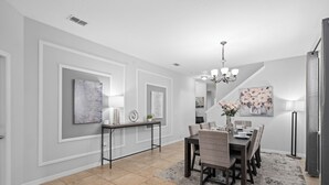 A dining area with a dark wooden table, beige chairs, and a gray rug. There is a chandelier above the table and artwork on the walls.