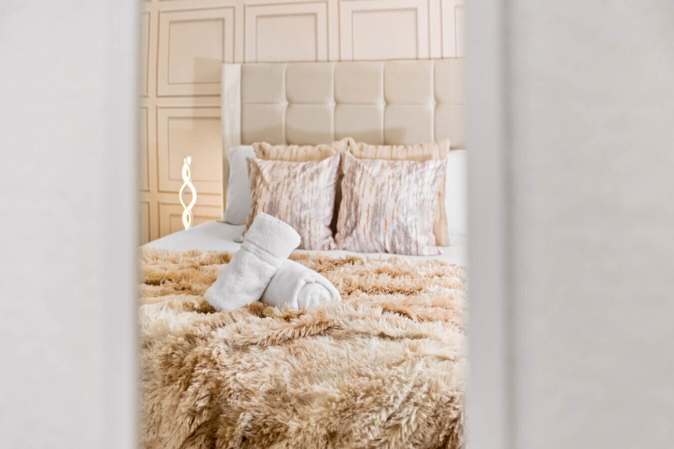 Ein ordentlich bezogenes Bett mit flauschigem, beige farbenen Bettzeug und einem weißen Handtuch, einem gepolsterten Kopfteil und einer dekorativen Wandverkleidung.