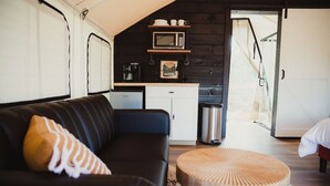 A cozy interior with a black leather sofa, a wooden coffee table, a kitchen area with a microwave and coffee maker, and a visible door leading to another room.