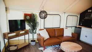 Ein gemütlicher Wohnbereich mit einem braunen Ledersofa, einem Holztisch, einem Fernseher auf einem Holzgestell, einer Topfpflanze und einer Küchenzeile mit Mikrowelle und Kühlschrank.