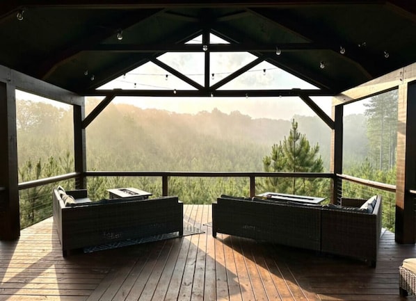A covered outdoor seating area with two cushioned benches and a table, overlooking a mountainous landscape.