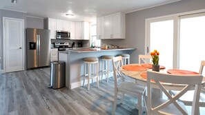 A modern kitchen with white cabinets, a stainless steel refrigerator, and a microwave. There is a dining area with a round table and chairs, and a trash can is visible. - Golden Tee in North Fort Myers (North Fort Myers)