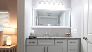 A bathroom with a white vanity, a mirror above the sink, and a lighted lamp on a bedside table. - Golden Tee in North Fort Myers (North Fort Myers)
