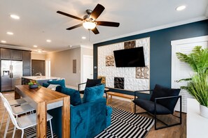 A modern living room with a dining area, a ceiling fan, a flat-screen TV mounted on a textured wall, and a potted plant.
