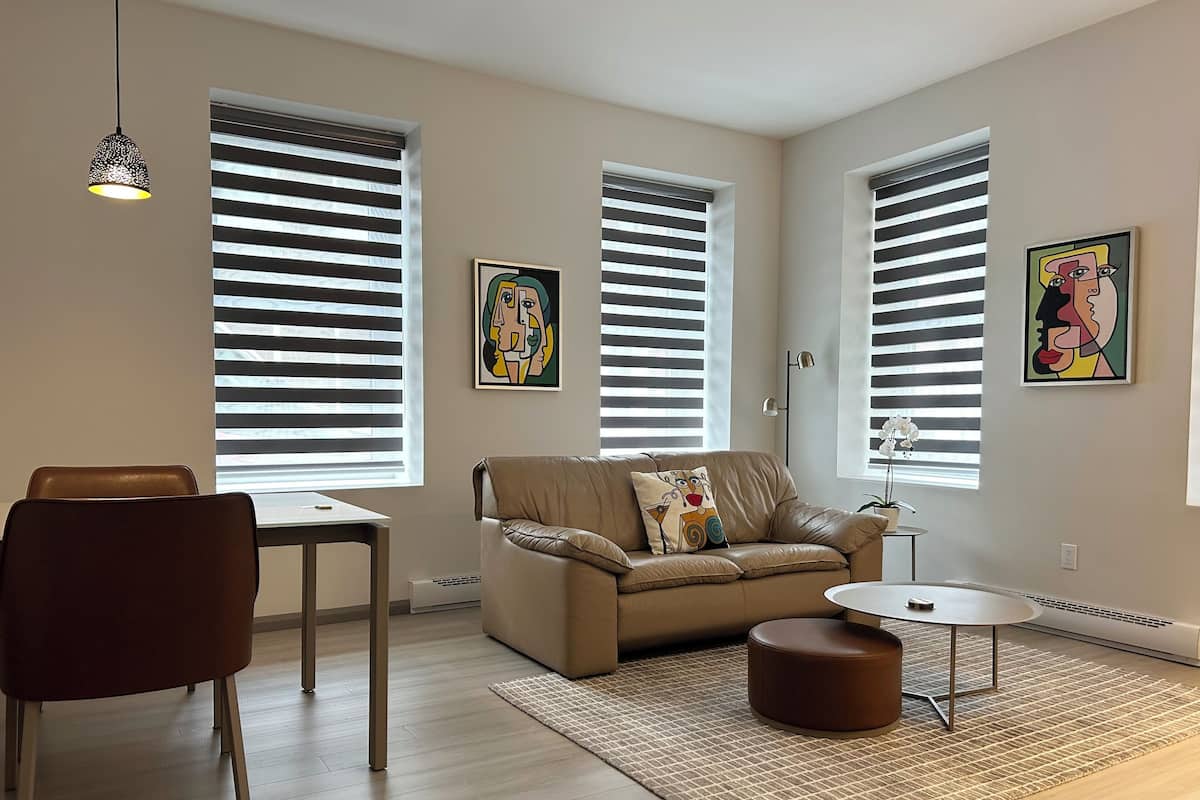 A modern living room with a sofa, a dining table, and a coffee table. There are two windows with blinds, a floor lamp, and two wall-mounted artworks.