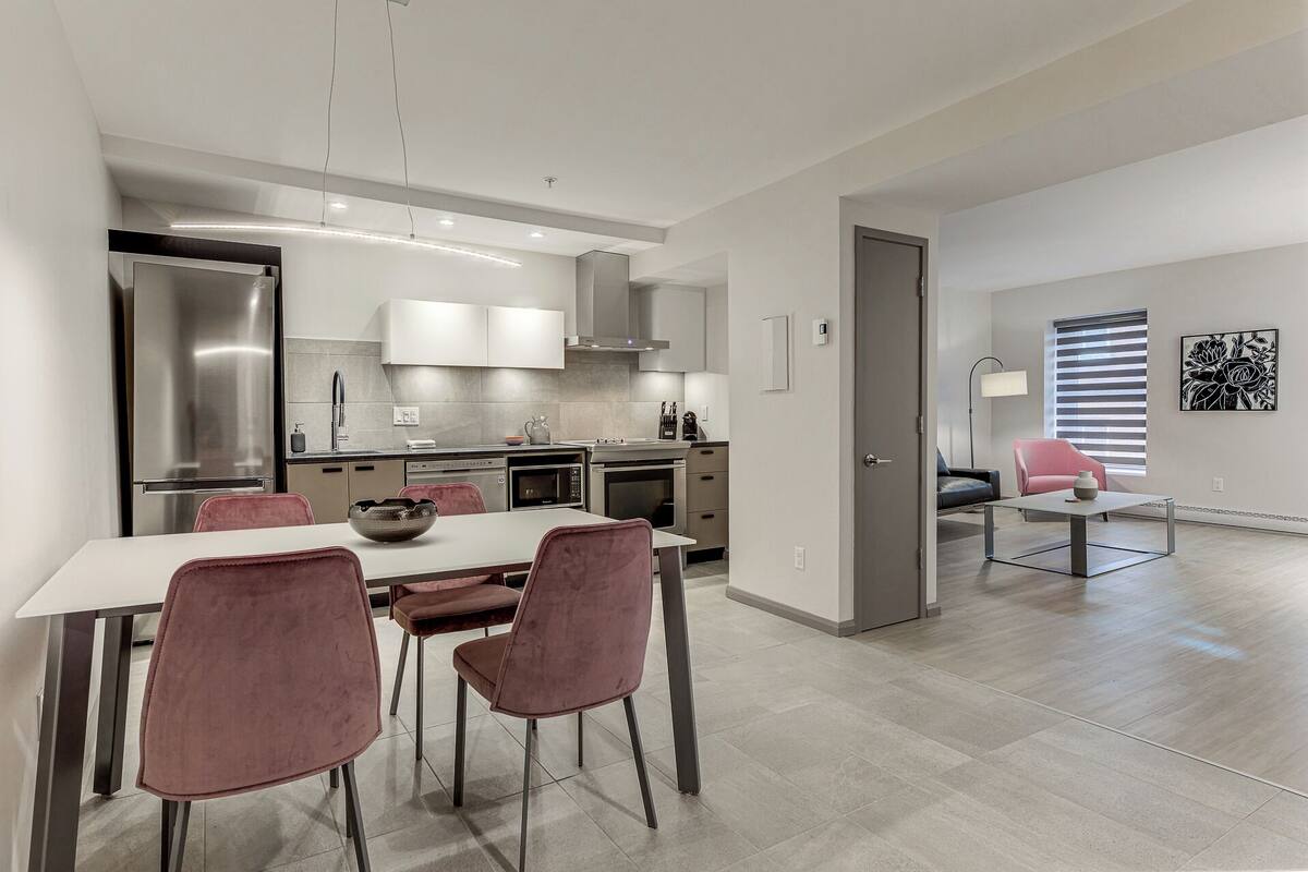 A modern kitchen with stainless steel appliances, a dining area with pink chairs, and a minimalist living space with a sofa and a coffee table.