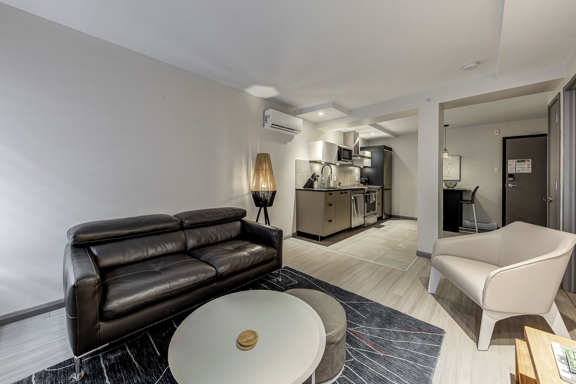 A modern living room with a black leather sofa, a white coffee table, and a beige armchair. A kitchen is visible in the background with stainless steel appliances.