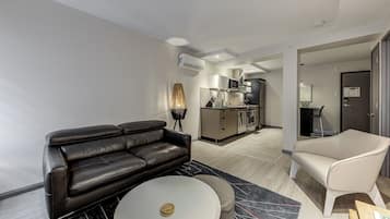 A modern living room with a black leather sofa, a white coffee table, and a beige armchair. A kitchen is visible in the background with stainless steel appliances.