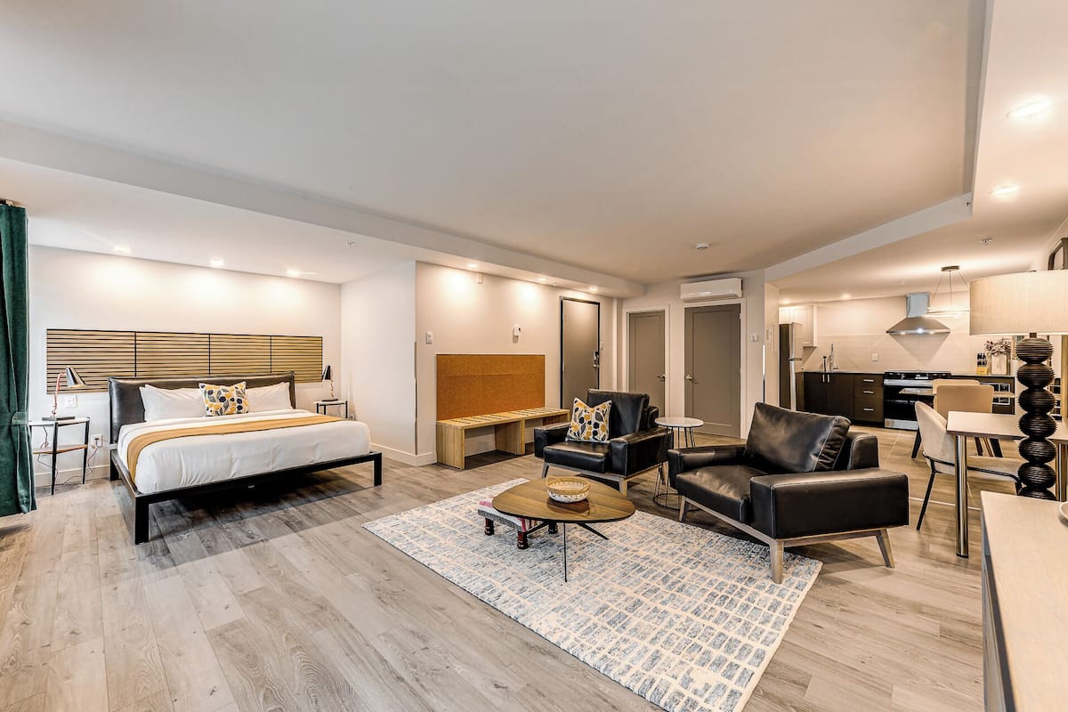 A modern hotel room with a large bed, a seating area with a coffee table, a small kitchen, and a separate dining area.