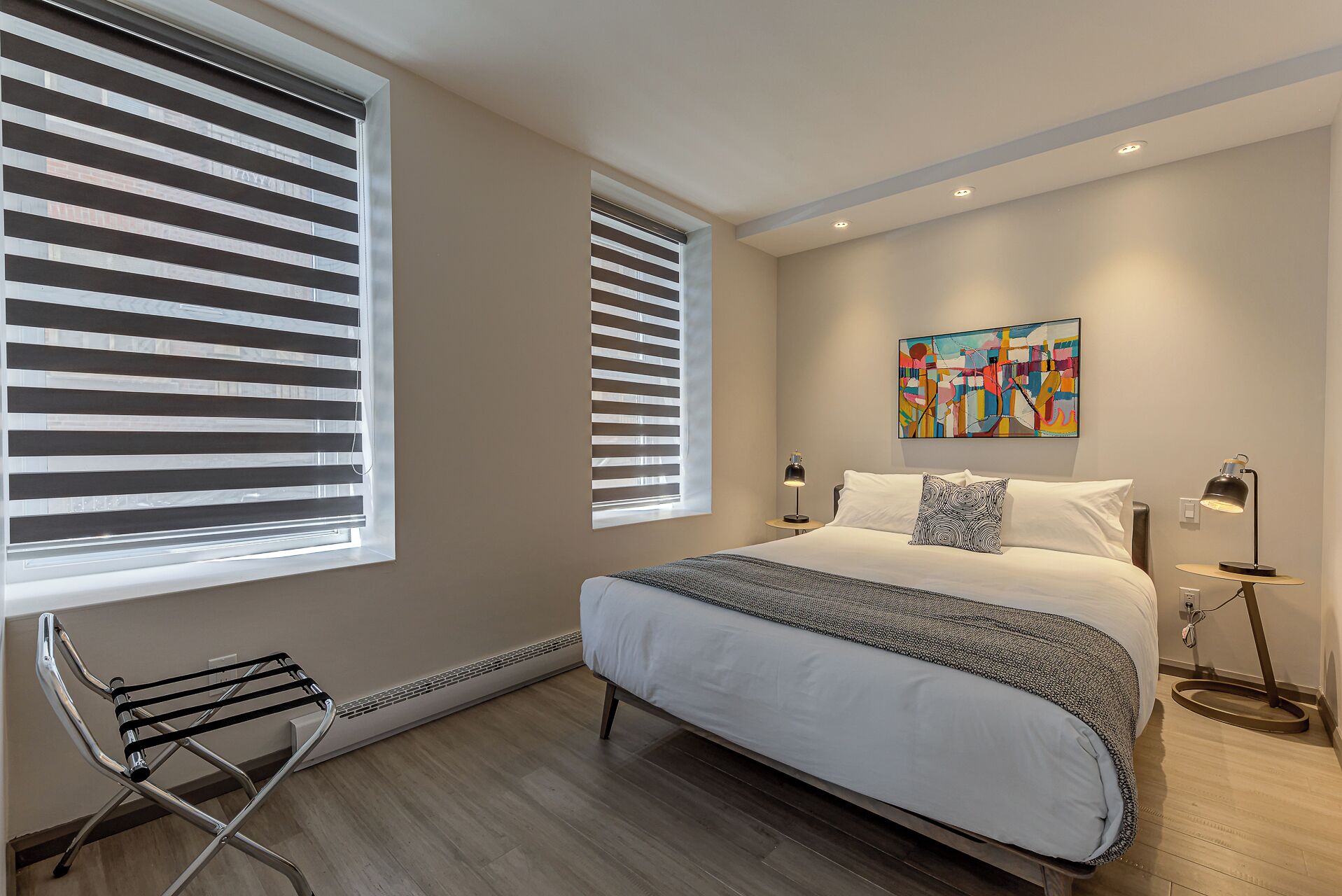 Un dormitorio con una cama, dos lámparas de noche, una mesita, una silla y un cuadro abstracto y colorido en la pared.