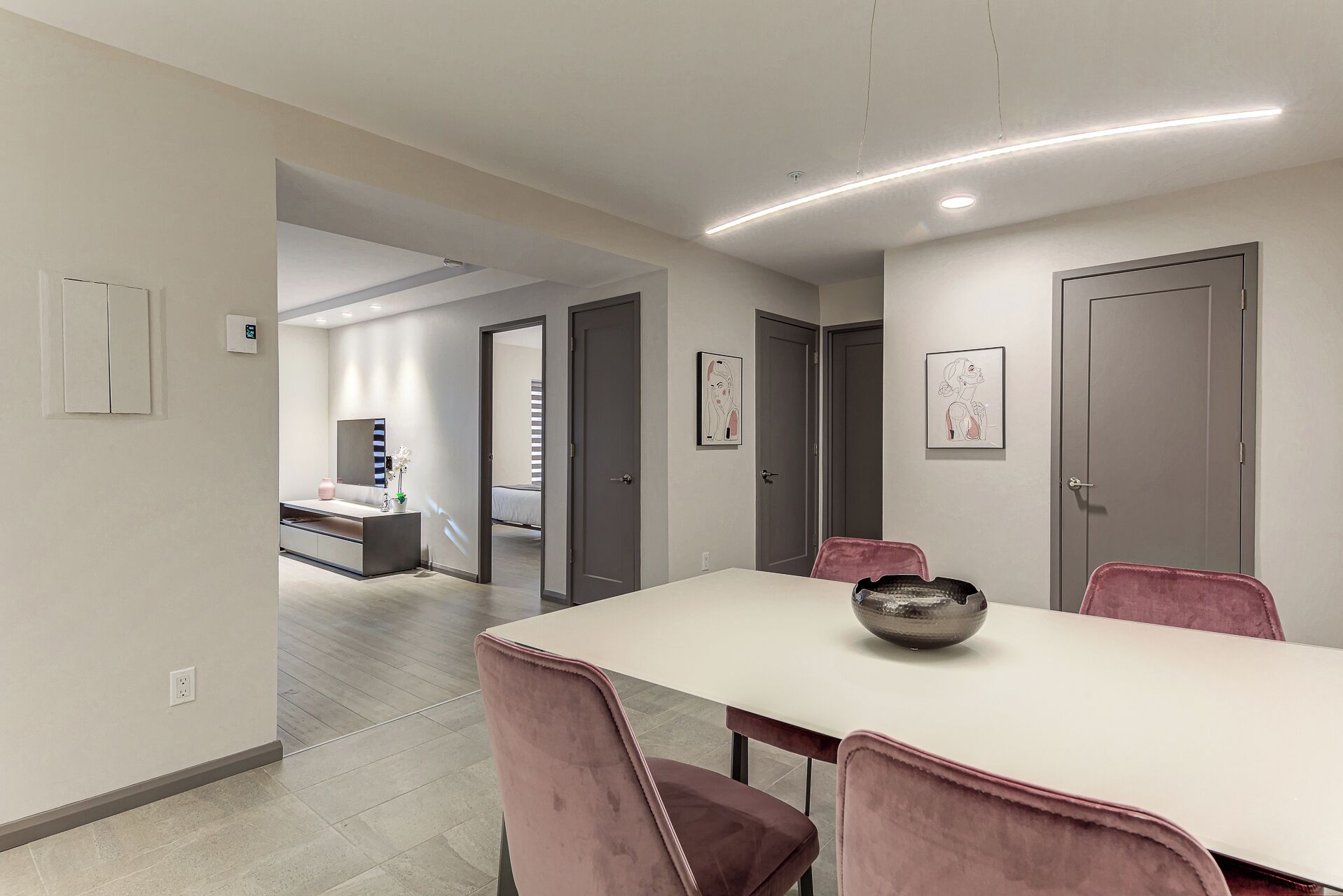 A modern dining area with a white table and pink chairs, a kitchen in the background, and artwork on the walls.
