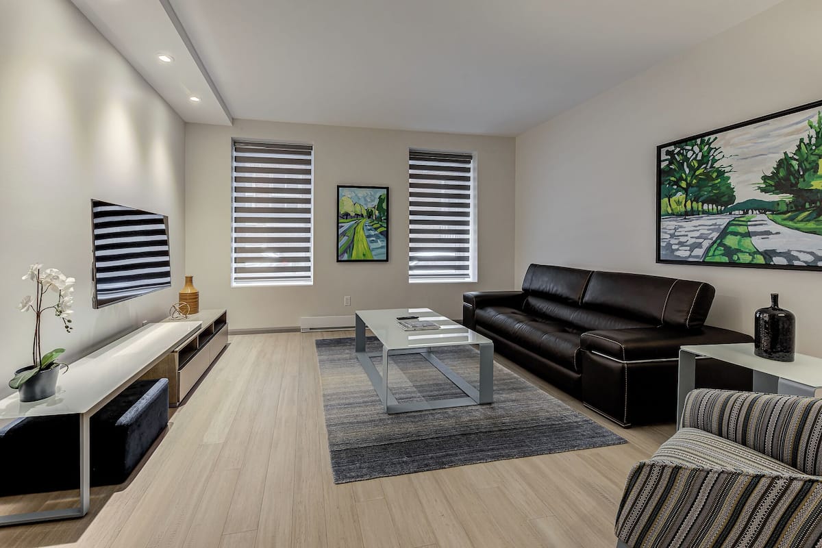 A modern living room with a black leather sofa, a coffee table, a TV mounted on the wall, and two windows with blinds.