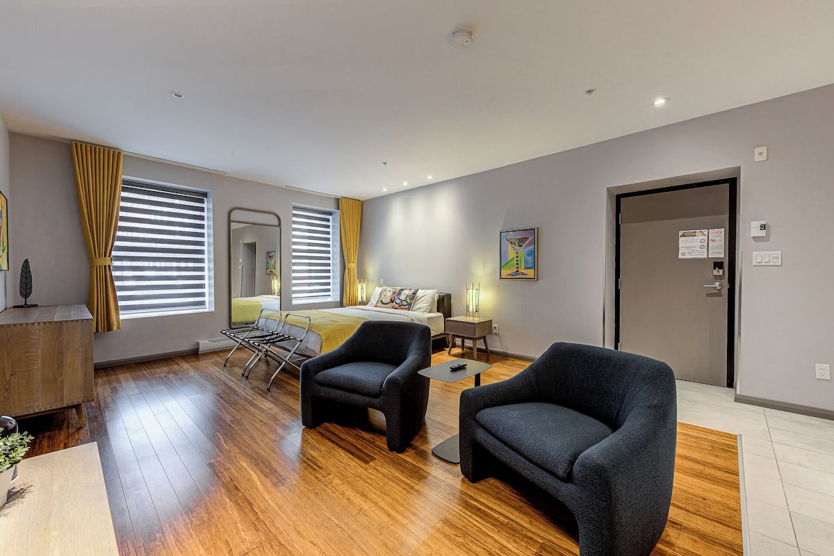 A modern hotel room with a bed, two armchairs, a small table, and a window with blinds.