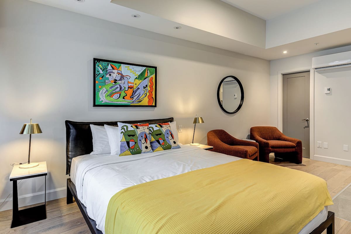 A modern bedroom with a bed, two chairs, a painting, and a mirror on the wall.