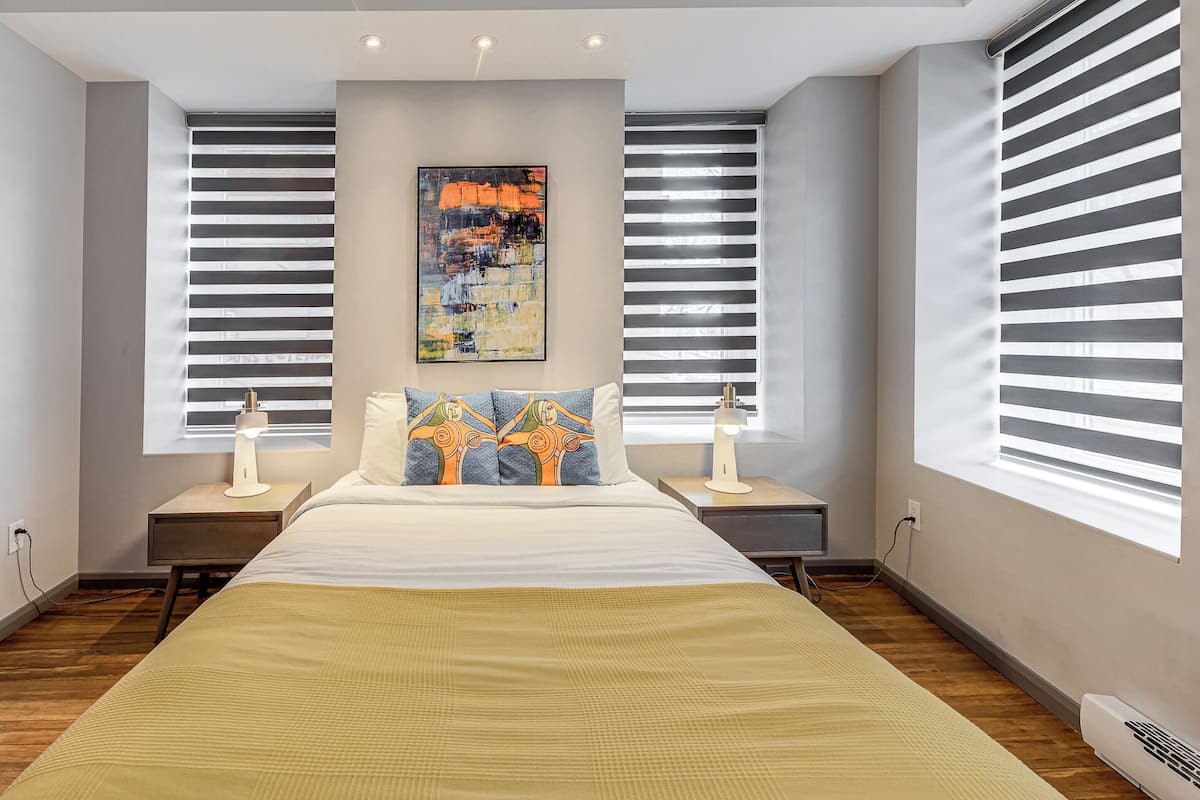 A modern bedroom with a large bed, two bedside tables, a wall-mounted painting, and large windows with blinds.