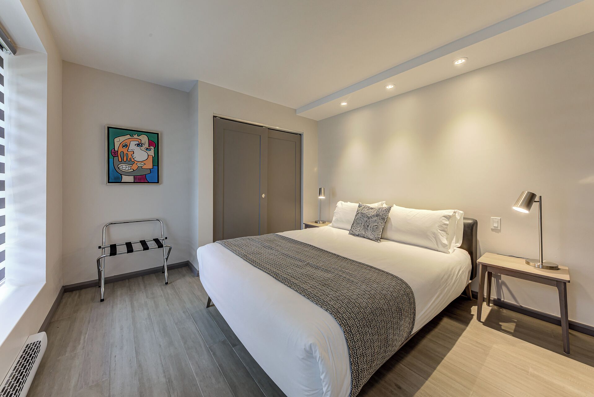 A modern hotel room with a large bed, a bedside table with a lamp, a wardrobe, and a radiator.