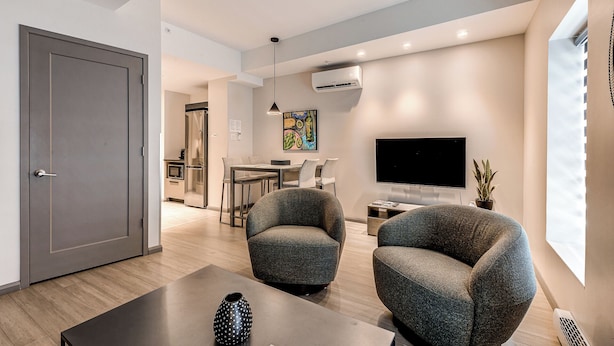 A modern living room with two armchairs, a coffee table, a flat-screen TV, and a dining area with a kitchen in the background. - 77 St-Vallier Appart Hôtel (Québec)