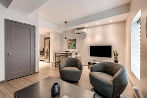 A modern living room with two armchairs, a coffee table, a flat-screen TV, and a dining area with a kitchen in the background. - 77 St-Vallier Appart Hôtel (Québec)