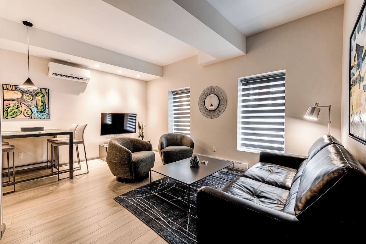 A modern living room with a leather sofa, two armchairs, a coffee table, and a TV. There is a dining area with a bar stool and a small table. The room has a large window with blinds, a ceiling fan, and a wall-mounted air conditioner.