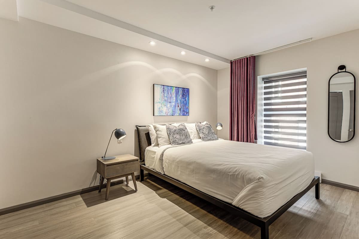A modern bedroom with a bed, bedside lamps, a nightstand, a painting, a window with blinds, and a mirror.