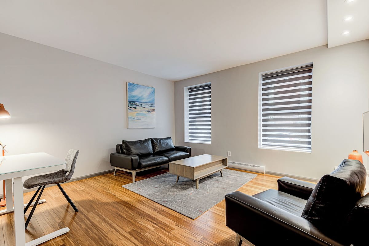 A modern living room with a black leather sofa, a glass coffee table, and a grey rug. There is a painting on the wall and two windows with blinds.