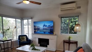 A living room with a fireplace, a television mounted above it, and a large window providing a view of trees. - Relax in a cozy house,BBQ,10 mins from the beaches (Miranda)