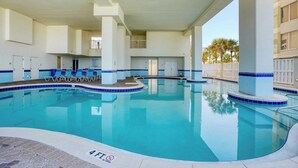 Une grande piscine intérieure à l’eau d’un bleu limpide, entourée de piliers blancs et bordée de carreaux bleus. Des chaises longues sont disposées le long du bord de la piscine, et une petite terrasse supérieure est aménagée.